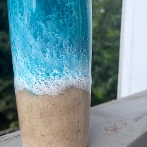 May include: A tall, cylindrical tumbler with a coastal theme. The top half features a vibrant blue and white wave design, transitioning to a sandy beige base. The tumbler is likely made of a durable material.