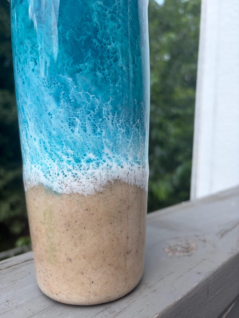 May include: A tall, cylindrical tumbler with a coastal theme. The top half features a vibrant blue and white wave design, transitioning to a sandy beige base. The tumbler is likely made of a durable material.