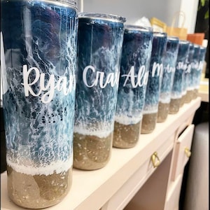 May include: A row of personalized tumblers with ocean-themed designs. The tumblers are clear with a blue and white ocean wave design and a brown sand bottom. The names "Ryan", "Craig", "Alo", "M", "Shay", and "Josh" are written on the tumblers.