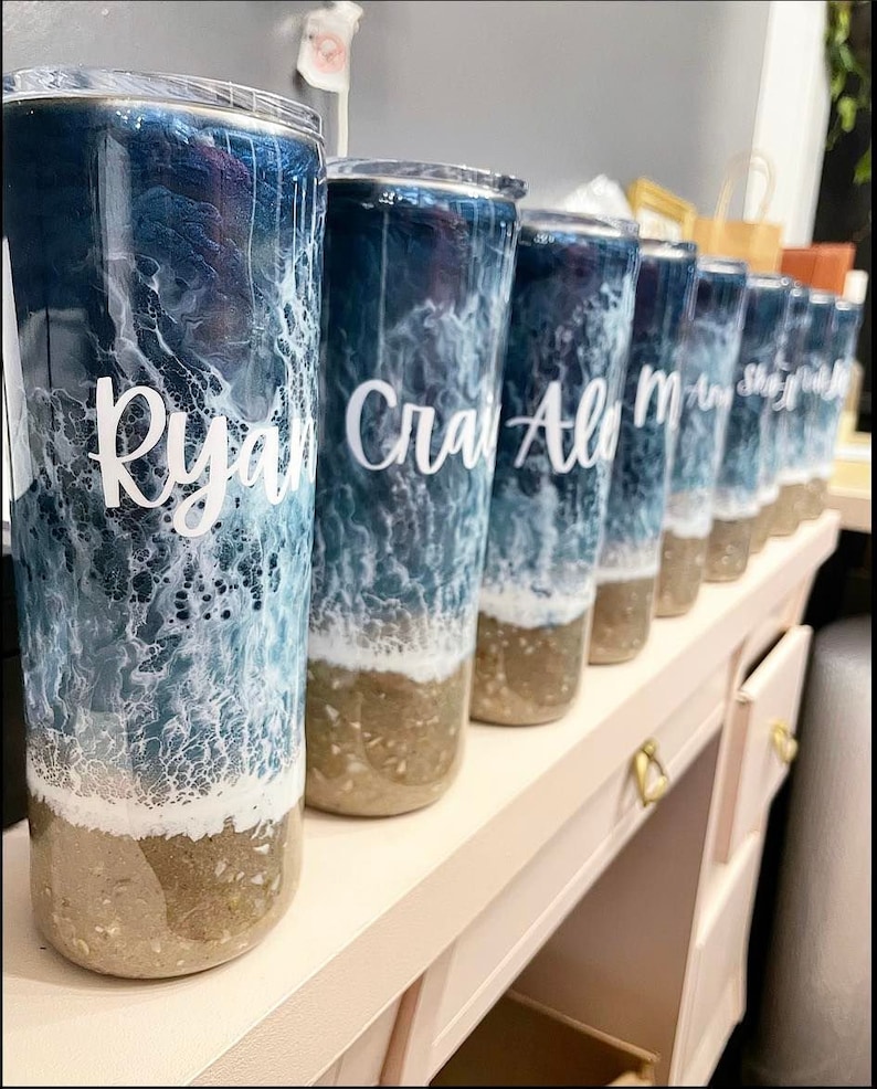 May include: A row of personalized tumblers with ocean-themed designs. The tumblers are clear with a blue and white ocean wave design and a brown sand bottom. The names "Ryan", "Craig", "Alo", "M", "Shay", and "Josh" are written on the tumblers.