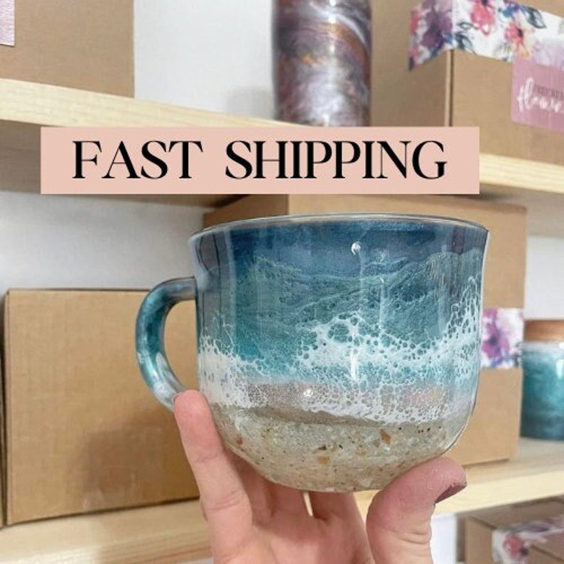 Peut inclure: Une tasse en c&eacute;ramique bleue et blanche avec un motif de plage. La tasse a un bord blanc et une anse. La sc&egrave;ne repr&eacute;sente de l'eau bleue, des vagues blanches et une plage de sable.