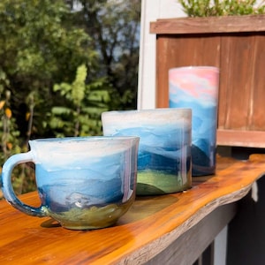 May include: Three ceramic mugs with a blue, green, and white watercolor design. The mugs are sitting on a wooden surface.