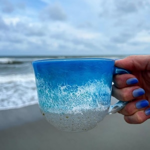 Peut inclure: Une tasse en c&eacute;ramique bleue et blanche avec un motif de vague. La tasse est remplie d'un liquide blanc et a un fond sablonneux.