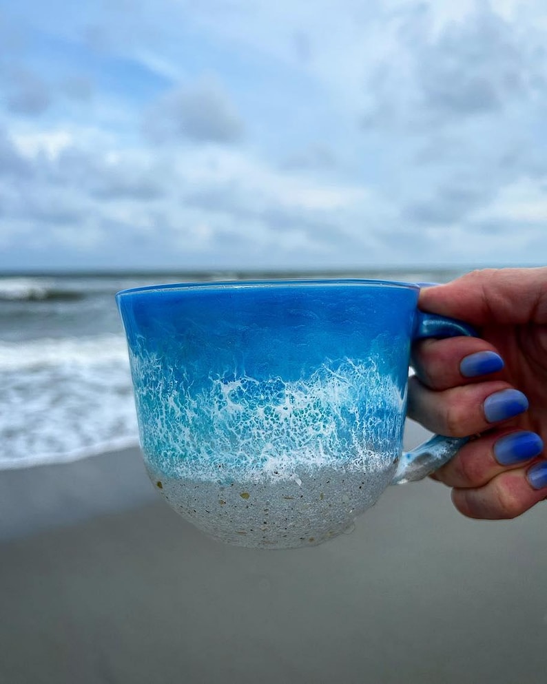 Peut inclure: Une tasse en c&eacute;ramique bleue et blanche avec un motif de vague. La tasse est remplie d'un liquide blanc et a un fond sablonneux.