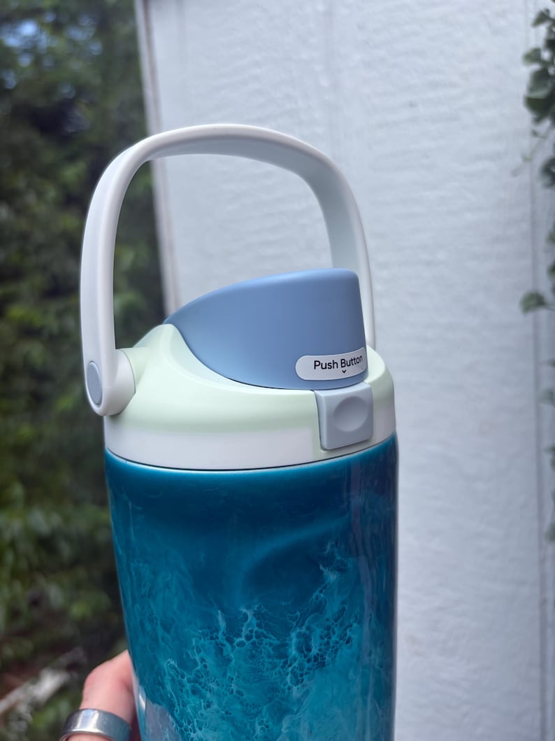 May include: A teal and white water bottle with a handle. The bottle has a push button on the lid and a white handle. The body of the bottle has a swirling pattern of teal and white. The lid is light blue and white.