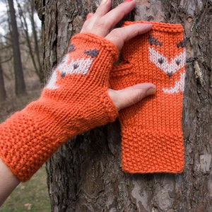 Puede incluir: Guantes sin dedos de punto naranja con un diseño de zorro blanco. Los guantes están doblados por la mitad y sostenidos en una mano.