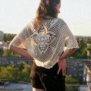 May include: Cream-colored, open-weave crochet shawl with a floral design on the back. The shawl has a square shape and short sleeves. The garment is worn over a dark top and black shorts.