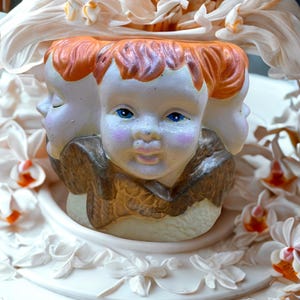 May include: A white ceramic sculpture featuring three cherubic faces with orange hair and blue eyes. The sculpture is adorned with white and orange orchids, creating a floral arrangement. The base is decorated with floral relief.