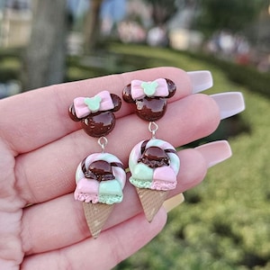 May include: A pair of whimsical dangle earrings. The earrings feature a brown Mickey Mouse head with a pink bow and a mint green bow accent. Below, a colorful ice cream cone design with pink, mint green, and chocolate-colored elements.