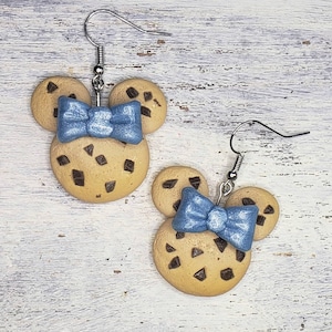 Könnte beinhalten: Ein Paar Ohrringe mit einem Schokoladenkeks-Design und einer blauen Schleife. Die Kekse sind braun mit Schokoladenstückchen und die Schleifen sind hellblau.