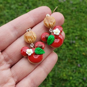 May include: A pair of dangle earrings featuring charms shaped like red glazed donuts with green leaves and small white flowers. Above each donut charm is a smaller tan circular piece with brown textured lines, all connected by silver metal hooks.