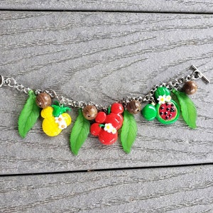 Op de afbeelding: Een zilveren armband met fruitvormige bedels, waaronder een ananas, een watermeloen en een aardbei. De bedels zijn bevestigd aan een zilveren ketting met bruine houten kralen.