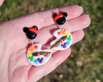 May include: A pair of colourful paint palette earrings with a Mickey Mouse head and a paintbrush charm. The palette is white with rainbow coloured paint splatters and a Mickey Mouse head in the centre.