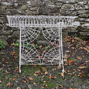 May include: White metal plant stand with a circular top and a spiderweb-like design. The stand is on a gravel surface with brown leaves.