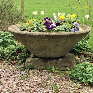 May include: A large, grey concrete planter with a wide, shallow bowl shape. The planter is filled with a variety of colourful flowers, including purple pansies, yellow daffodils, and white flowers. The planter is sitting on a stone base and is surrounded by gravel.