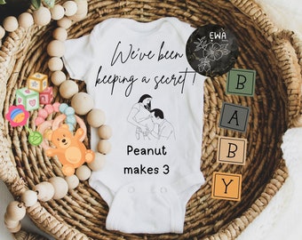 May include: White onesie with the text "We've been keeping a secret!" and a line drawing of a couple. Below the drawing is the text "Peanut makes 3". The onesie is in a wicker basket with wooden beads and toy blocks.