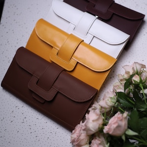 Pode incluir: Quatro carteiras de couro em marrom, amarelo, branco e marrom escuro. As carteiras estão empilhadas uma em cima da outra, com a carteira marrom na parte inferior e a carteira marrom escuro na parte superior. As carteiras têm um design simples com fecho de aba e uma única alça.