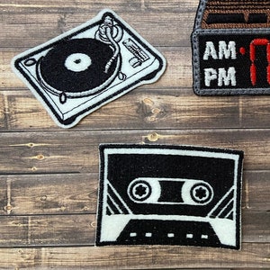 Könnte beinhalten: Drei gestickte Aufnäher mit einem Plattenspieler, einem Kassettenband und einer Uhr mit AM- und PM-Anzeige. Die Aufnäher sind weiß mit schwarzen und grauen Stickereien.