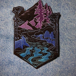 以下が含まれることがあります： 濃い茶色の背景に、紫色の山とピンク色の木々のある風景の中を蛇行する青い川が描かれた刺繍パッチです。