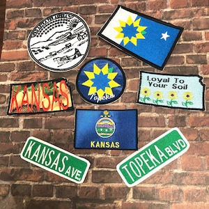 Könnte beinhalten: Eine Sammlung gestickter Aufnäher mit Symbolen und Sehenswürdigkeiten des Bundesstaates Kansas. Die Aufnäher beinhalten ein Staatssiegel, eine Flagge, ein Straßenschild und ein Design mit Sonnenblumen. Der Text auf den Aufnähern lautet "Kansas", "Topeka", "Kansas Ave", "Topeka Blvd" und "Loyal To Your Soil".