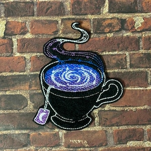 May include: An embroidered patch of a black teacup with a swirling purple and blue design inside. The teacup has a white string with a purple tea bag that has a crescent moon on it. The teacup has a white steam coming from it.