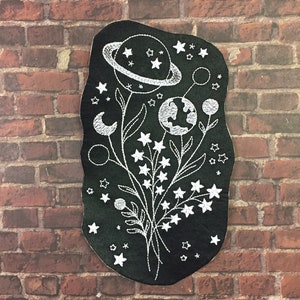 May include: A black embroidered patch with a white outline of a bouquet of flowers with planets, stars, and a crescent moon. The flowers are stylized and have a whimsical feel.