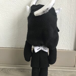 May include: Black plush toy with a white bow tie and a white headband. The toy is standing on a wooden floor.
