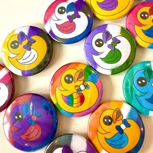 Könnte beinhalten: Eine Sammlung bunter, runder Buttons mit einem Cartoon-Enten-Design. Jeder Button hat ein anderes Farbschema, darunter Regenbogen, Gelb, Lila, Blau und Grün. Die Enten haben eine Fliege und ein lächelndes Gesicht.