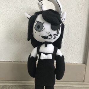 May include: Black and white plush toy with a black dress, a bow, and a single eye. The toy has a split face with one side showing a cartoonish smile and the other side showing a more sinister expression.
