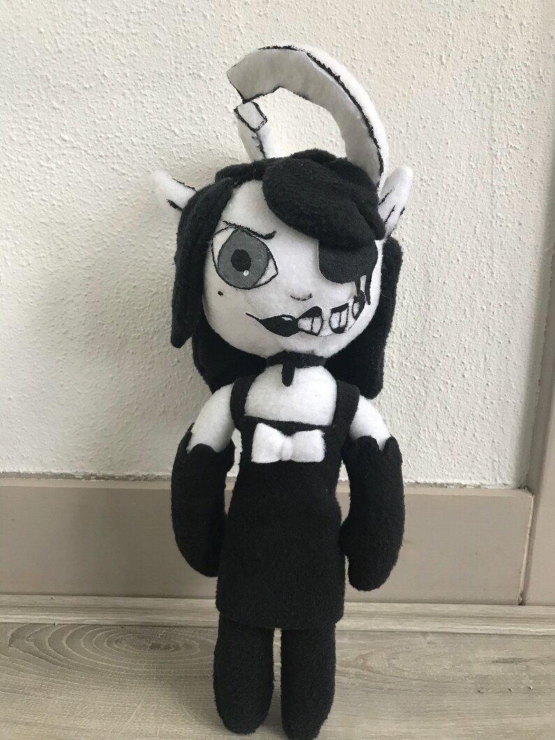 May include: Black and white plush toy with a black dress, a bow, and a single eye. The toy has a split face with one side showing a cartoonish smile and the other side showing a more sinister expression.