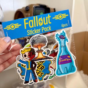 Peut inclure: Un paquet de 6 autocollants Fallout dans une pochette en plastique transparent avec une carte d'en-tête bleue. Les autocollants représentent un personnage en combinaison bleue, un champignon atomique et une bouteille bleue Quantum.