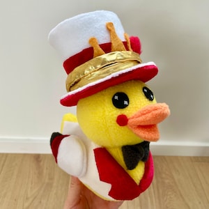 Op de afbeelding: Een gele pluche eend met een rood-witte hoge hoed met een gouden band en een zwarte strik.