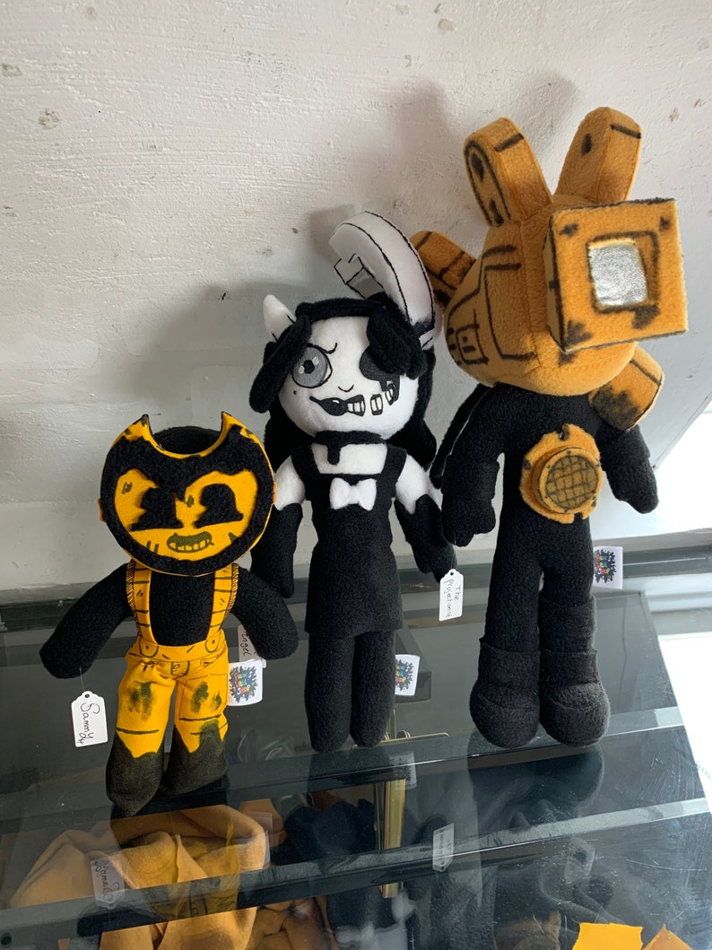 May include: Three plush toys from the video game Bendy and the Ink Machine. The toys are a black and white character with a bow tie, a yellow and black character with a large head, and a yellow and black character with a large head and a camera.