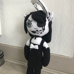 May include: Black and white plush toy with a black dress, white bow, and black gloves. The toy has a black and white face with a single eye and a dripping black substance.