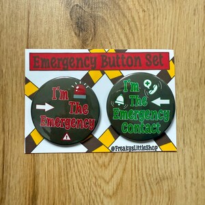 Op de afbeelding: Een set van twee ronde buttons op een witte kaart met een bruin, geel en zwart ontwerp. Eén button leest "I'm The Emergency" met een rode sirene-afbeelding. De andere leest "I'm The Emergency Contact" met een groene bel-afbeelding.