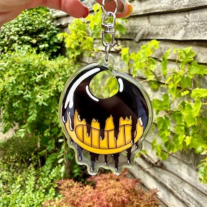 Op de afbeelding: Een doorzichtige acryl sleutelhanger met een cartoongezicht met een brede gele grijns en zwarte inkt die van bovenaf naar beneden druipt. Het ontwerp is ingesloten in transparant materiaal met een zilverkleurige metalen karabijnhaak.