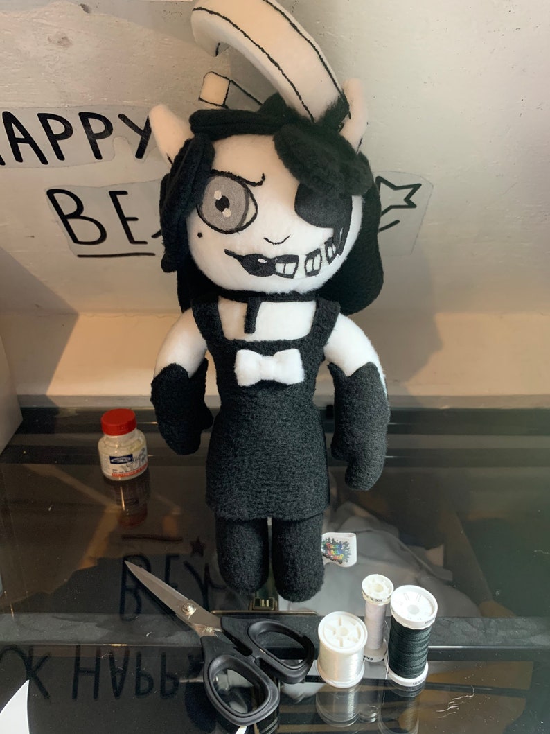 May include: Black and white plush toy with a white eye patch, black hair, and a black dress. The toy is standing on a table with scissors, white and black thread, and a bottle of glue.