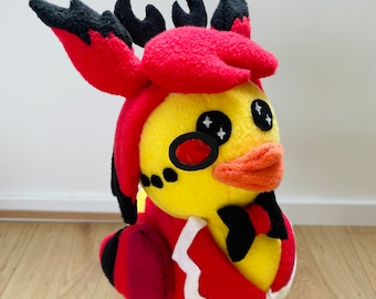 Puede incluir: Un pato de peluche con cuernos rojos y negros, una chaqueta roja y blanca y una pajarita negra. El pato tiene plumas amarillas, un pico rojo y ojos negros con destellos blancos.