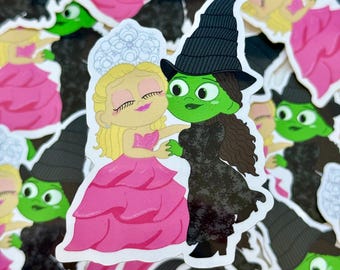 Op de afbeelding: Een sticker met een cartoonillustratie van twee vrouwen. Een vrouw draagt een roze jurk en een witte tiara. De andere vrouw draagt een zwarte heksenhoed en een zwarte jurk. De vrouwen knuffelen elkaar.