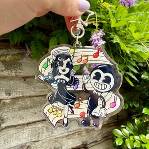 Op de afbeelding: Een doorzichtige acryl sleutelhanger met een cartoonillustratie van twee zwart-witte figuren die dansen op een notenbalk met kleurrijke muzieknoten. De sleutelhanger heeft een zilverkleurige, stervormige metalen sluiting aan de bovenkant.