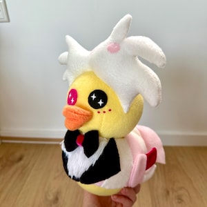 Op de afbeelding: Een gele pluche eend met wit haar, roze wangen en een zwarte strik. De eend draagt een roze en witte jurk.