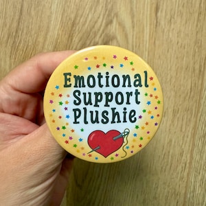 Op de afbeelding: Een ronde, gele button met de tekst "Emotional Support Plushie" in het groen. De button heeft een rood hart doorboord met een naald en draad, omringd door kleurrijke sterren.