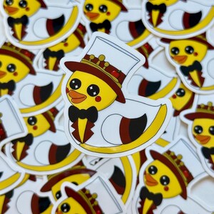 Op de afbeelding: Een stapel gele rubberen eendenstickers met een witte hoge hoed, een rode strik en een kroon. De eenden hebben zwarte ogen en een rode snavel.