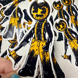 以下が含まれることがあります： 丸い黒い頭、大きな笑顔、黄色い目をした漫画のキャラクターが描かれたダイカットステッカー。キャラクターは黒いインクのような滴が垂れた黄色いオーバーオールを着ており、片手に黄色い頭の斧を持っています。