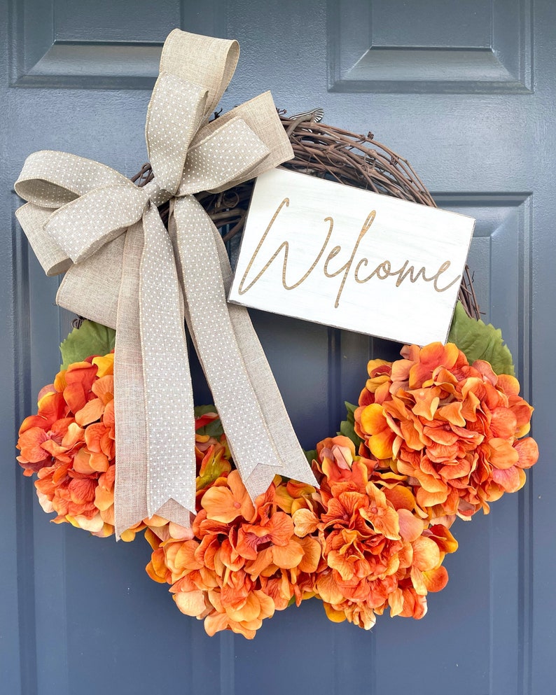 May include: A fall wreath with orange hydrangeas and a brown burlap bow with white polka dots. The wreath has a wooden sign that says 'Welcome' in gold lettering.