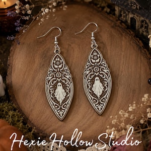 May include: A pair of dangle earrings featuring a pointed, leaf-shaped wooden design with an intricate, symmetrical white filigree pattern and a central ghost illustration. The earrings are suspended from silver-toned metal hooks.