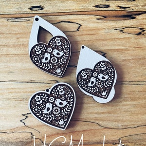 May include: Three wooden heart-shaped ornaments with a white background and brown floral and bird designs. The ornaments are on a wooden surface with the text "HG Market 2022" written in white.