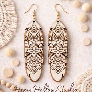 May include: Pair of wooden feather earrings with intricate mandala designs, featuring gold-colored hooks. The earrings are light brown with a cream-colored background and the text "Hexie Hollow Studio".