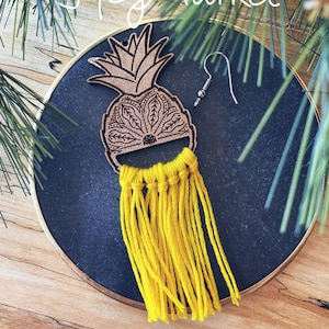May include: A pair of yellow tassel earrings with a wooden pineapple design. The earrings are on a black plate with a gold rim. The text "HG Market" is visible in the background.