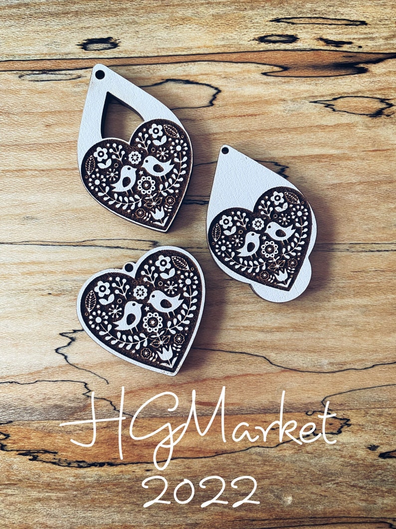 May include: Three wooden heart-shaped ornaments with a white background and brown floral and bird designs. The ornaments are on a wooden surface with the text "HG Market 2022" written in white.